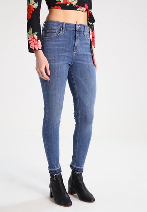 Femme portant un jean skinny bleu taille haute avec des ourlets effilochés, des bottines noires et un top court noir à manches longues à motif floral.