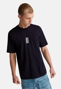 Mladý muž v černém tričku s bílým nápisem "OUT", dívající se dolů, v kombinaci se světlými modrými džíny na jednobarevném bílém pozadí.