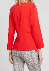 Rød langærmet top med en tætsiddende talje og en lynlås på ryggen, parret med grå slangeskindsprintbukser med en glat tekstur og design.