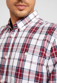 Uomo che indossa una camicia a quadri rossa, bianca e blu con taschino frontale e colletto abbottonato, mostrando la parte bassa del viso e il collo.