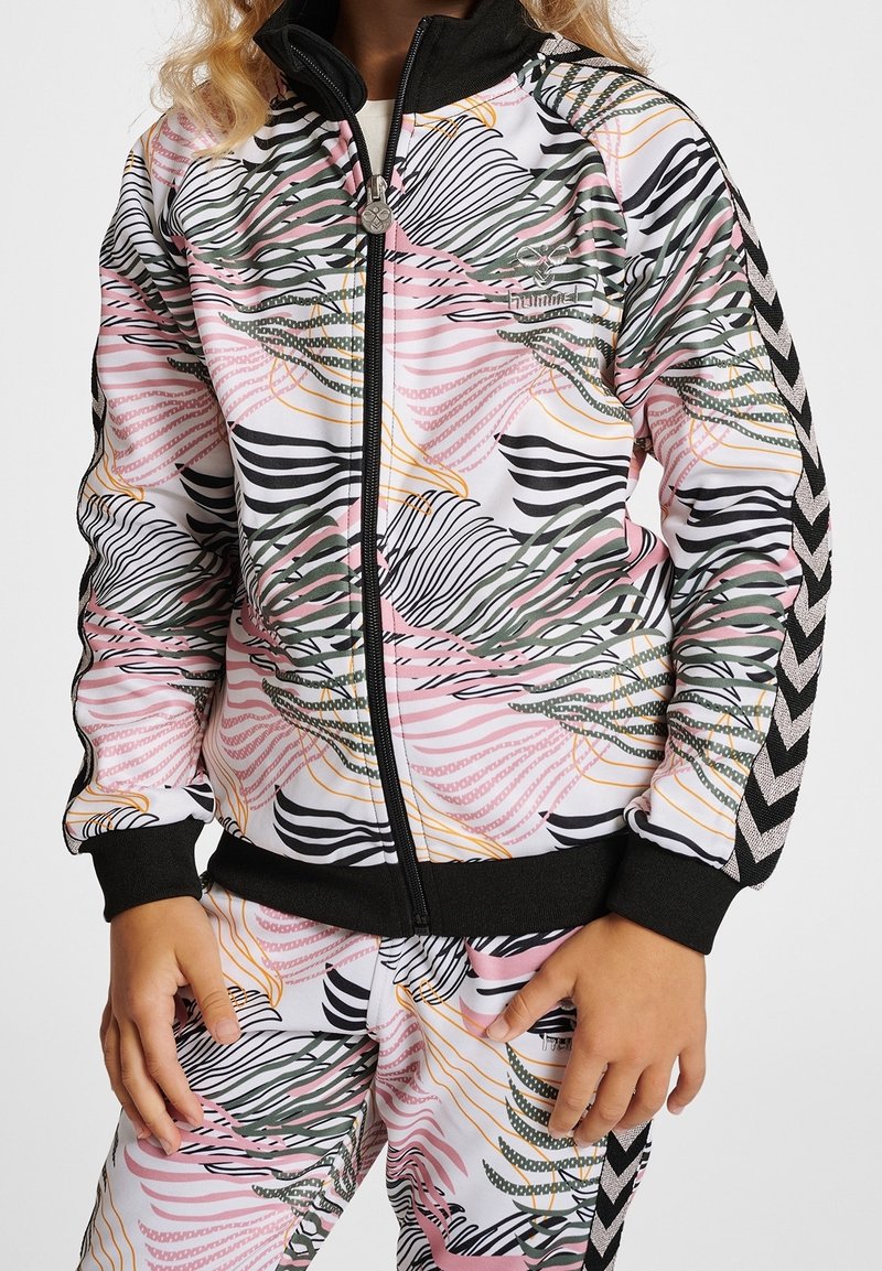 Kind, das einen bunten, gemusterten Trainingsanzug mit schwarzen Bündchen und Zickzackstreifen an Ärmeln und Hosen trägt, steht mit entspannten Händen.