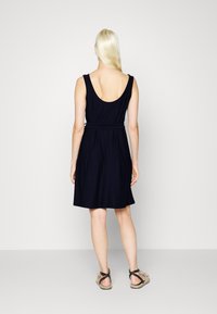 Vestido sem mangas azul-marinho com costas baixas, faixa na cintura e saia evasê. Combinado com sandálias bege com tiras entrelaçadas. Textura suave.