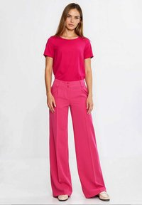 Różowy top z krótkim rękawem w zestawieniu z różowymi, szerokimi spodniami z wysokim stanem, posiadającymi dwa widoczne guziki na talii, oraz płaskimi butami.