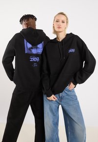Black hoodies with front pockets; one has a blue graphic on the back featuring a vinyl record and text. Models wear loose denim pants.