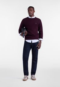 Maglione bordeaux, camicia bianca con colletto, jeans blu scuro, con una borsa nera. Sneakers con dettagli grigi e bianchi. Design semplice e moderno.