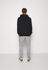 Mężczyzna stojący tyłem, ubrany w czarną bluzę z kapturem, szare spodnie dresowe cargo i kolorowe sneakersy na jednolitym jasnym tle.