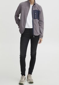 Giacchetto in pile grigio chiaro con tasca sul petto blu navy, chiusura con zip e colletto rialzato. Abbinato a jeans skinny neri e sneaker grigie.