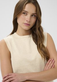 Smėlio spalvos be rankovėmis palaidinė iš minkštos, tekstūruotos medžiagos. Pasirašyta klasikinė apvalios iškirptės ir prigludusių siluetas. Matomos subtilios toninės variacijos.