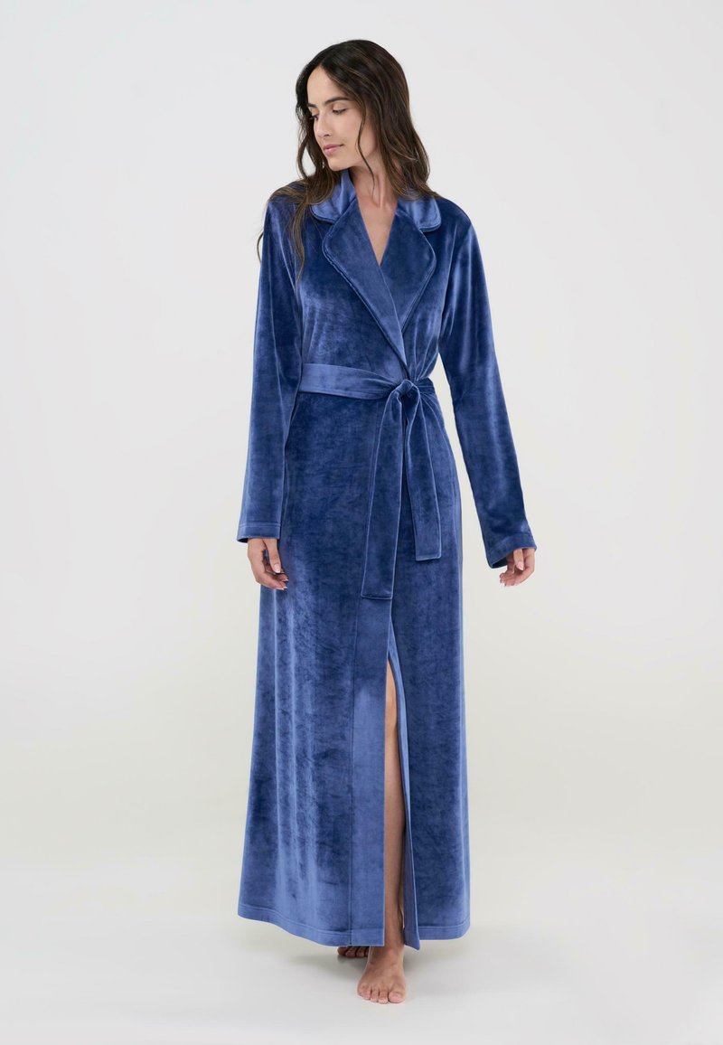 Larga bata de terciopelo azul con cuello de chal, diseño envolvente, cinturilla anudada y abertura lateral. Textura suave con mangas largas.