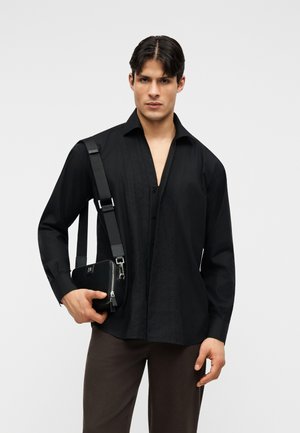 Άνδρας φορώντας μαύρο πουκάμισο με κουμπιά και καφέ παντελόνι, κρατώντας μια μαύρη τσάντα χιαστί με παχύ ρυθμιζόμενο ιμάντα στον ώμο του.