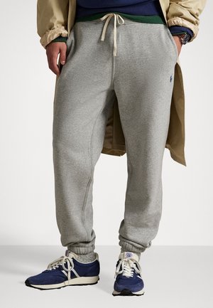 Mand iført lysegrå joggingbukser med snøre, beige trenchcoat, mørkeblå sneakers med hvide snørebånd og grå sokker, stående med hænderne i lommerne.