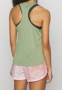 Top verde smanicato con rifiniture nere, caratterizzato da una texture liscia. Indossato con pantaloni corti rosa chiaro che presentano un sottile motivo tie-dye.