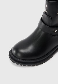 Bota negra impermeable con acabado brillante, punta redondeada, tobillo acolchado y suela de goma con patrones de tracción. Detalles de hardware de acento plateado.