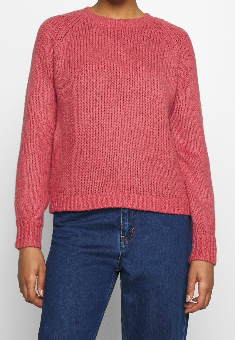 Růžový pletený svetr s kulatým výstřihem, dlouhými rukávy a žebrovanými lemy na rukávech a spodním okraji. Textura je robustní s viditelnými vzory pletení.