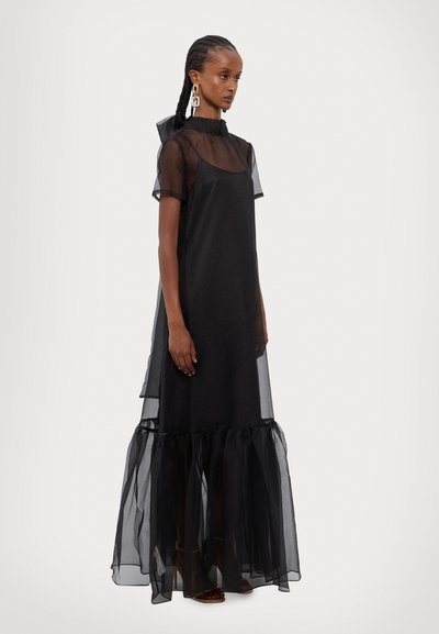 Rochie lungă neagră, transparentă, cu tiv cu volane și mâneci scurte. Are un guler înalt și un design stratificat, oferind o siluetă fluidă.