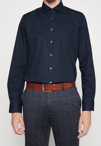 Marineblauw overhemd met lange mouwen en een klassieke kraag, vooraan knopen en een soepel fabric, gecombineerd met grijs geruite broek en een bruine leren riem.