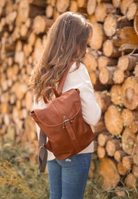 Zaino in pelle marrone con chiusura a patta, dettaglio con fibbia e spallacci regolabili; texture liscia su uno sfondo di tronchi impilati.