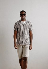 Camisa de manga curta com botões, com um padrão circular em bege e castanho, combinada com calções de cor clara. O modelo usa óculos de sol.