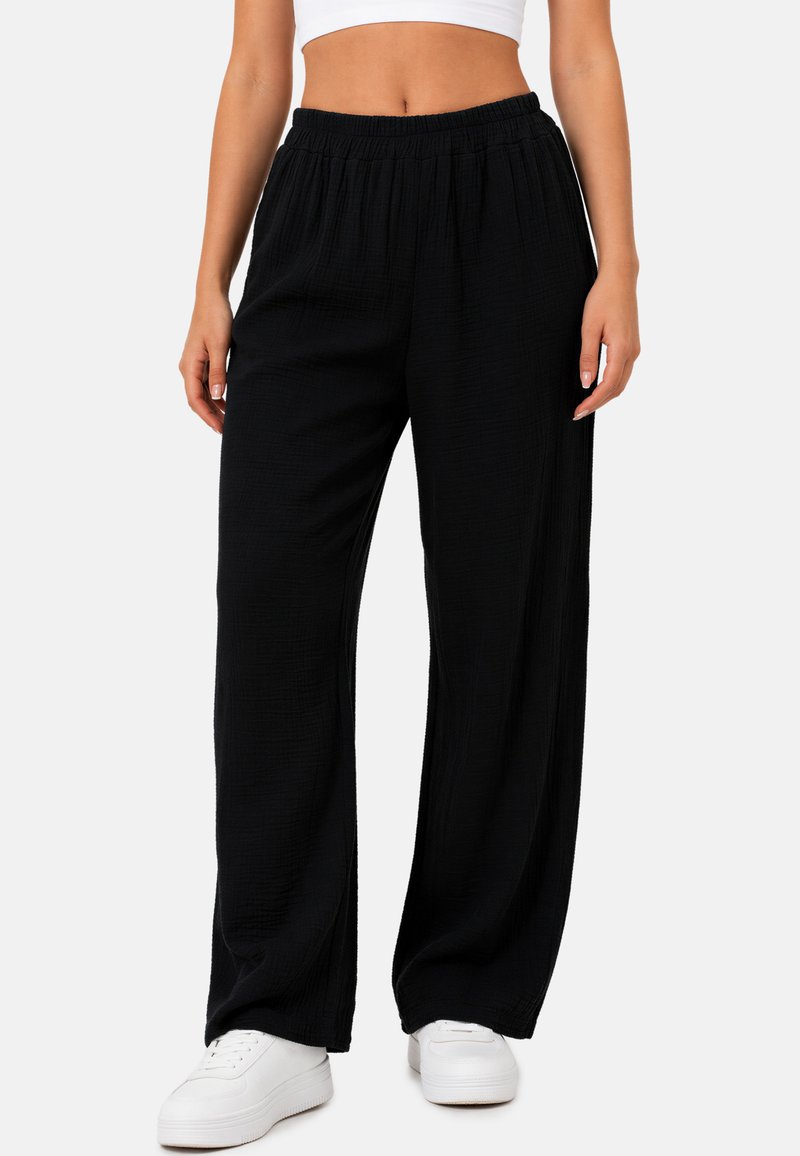 Femme portant un pantalon noir ample avec taille élastique et des baskets blanches, debout devant un fond clair uni.