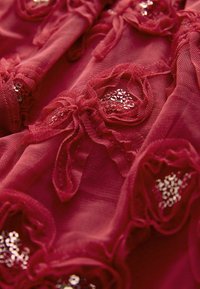 Röd tyg med texturerade rosett-mönster och glittrande paljetter inbäddade i centrum av varje rosett.