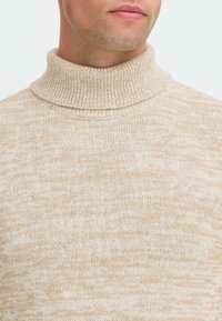Hombre vistiendo un suéter de cuello alto de punto jaspeado en beige y blanco, mostrando el cuello y la parte superior del pecho.