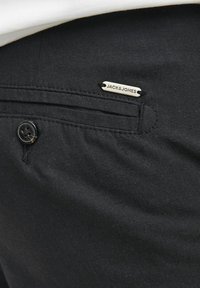 Crne hlače glatke teksture, sa prednjim džepom s srebrnom metalnom oznakom na kojoj piše "JACK & JONES" i kopčanjem na dugme.