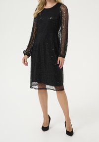 Robe en dentelle noire avec manches longues transparentes, présentant un motif texturé et des paillettes. Longueur genou avec un col rond. Portée avec des talons noirs.