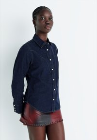 Ciemnobłękitna koszula z guzikami z klasycznym kołnierzykiem i białymi guzikami, w połączeniu z czerwono-czarną mini spódnicą ze skóry z dodatkowymi otworami.