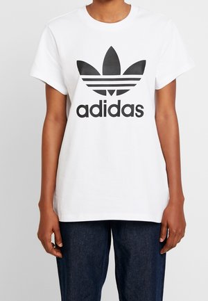 Osoba nosząca białą koszulkę Adidas z dużym czarnym logo trefoil i tekstem, zestawioną z ciemnoniebieskimi dżinsami.