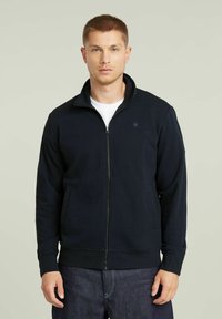 Giacca zip-up navy in tessuto morbido, con colletto alto, tasche laterali e polsini a costine per una vestibilità aderente.
