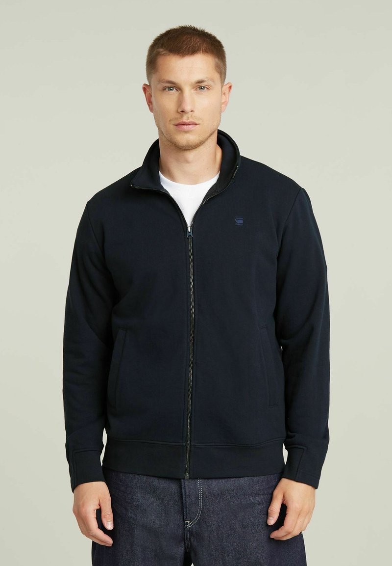 Giacca zip-up navy in tessuto morbido, con colletto alto, tasche laterali e polsini a costine per una vestibilità aderente.