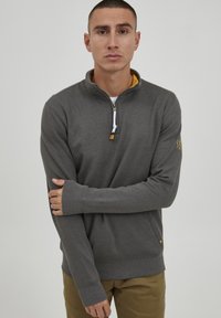 Sudadera gris con media cremallera en tejido suave, de manga larga, que presenta un interior del cuello amarillo, cremallera blanca y un pequeño logotipo en la manga.