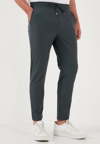 Donkergrijze joggingbroek van zacht materiaal, met een elastische tailleband met aantrekkoord en zijzakken. Gecombineerd met witte sneakers.