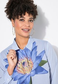 Camicia a righe blu e bianche con una grande grafica di un colorato fiore di loto blu sul petto, col collo a camicia e plastron con bottoni.