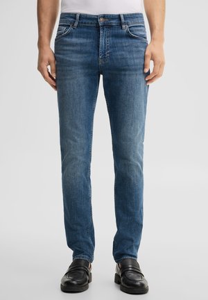 Uomo con jeans skinny blu, mocassini in pelle nera e top bianco, in piedi davanti a uno sfondo grigio chiaro uniforme.