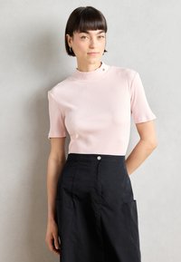 Helles rosa, geripptes Kurzarmoberteil mit einem hohen Kragen, das eine subtile Textur aufweist. Das Model kombiniert es mit einem dunklen Rock und Taschen.
