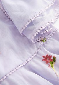 Helles lila, transparentes Gewebe mit gestickten rosa Blumen und grünen Akzenten, versehen mit einem zarten Pom-Pom-Bordür entlang der Kanten.