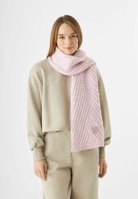 Jonge vrouw in een beige set van bijpassende sweatpants en een dikke, geribde roze sjaal om haar nek, met een witte achtergrond.