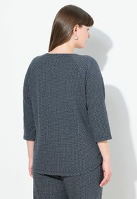Una donna con lunghi capelli castani indossa un top a maniche lunghe grigio scuro con un motivo e pantaloni coordinati, fronteggiando un muro bianco e proiettando un'ombra.