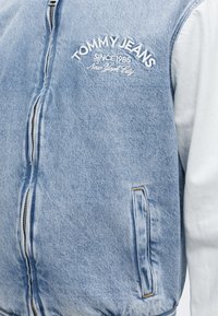Φωτεινό μπλε τζιν μπουφάν με λευκά μανίκια, φερμουάρ μπροστά, πλαϊνή τσέπη και κέντημα στο στήθος "TOMMY JEANS Since 1985 New York City".