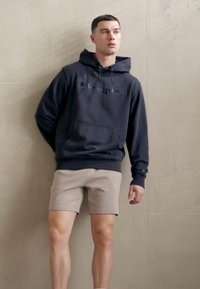 Haut à capuche marine avec une poche frontale et un cordon de serrage, arborant le logo "Champion". Associé à un short marron clair, sur fond neutre.