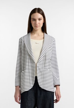 Vrouw met een wit colbert met zwarte horizontale strepen, een crèmekleurige geribbelde top en donkere wijd uitlopende jeans, staand tegen een witte achtergrond.