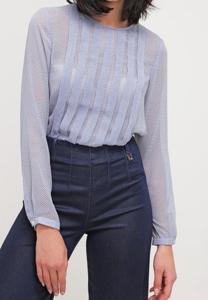 Femme portant un chemisier bleu clair à manches longues texturé, rentré dans un pantalon taille haute bleu foncé, sur un fond uni.