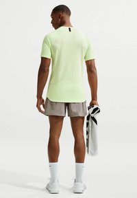 Άνδρας με ανοιχτό πράσινο αθλητικό πουκάμισο και γκρι σορτς κρατάει μαύρη και λευκή πετσέτα, όρθιος με την πλάτη προς τα εμπρός σε λευκό φόντο.