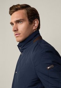 Giacca trapuntata blu navy con colletto alto, caratterizzata da dettagli in pelle marrone sulle spalle e una chiusura a scatto frontale. Texture liscia e design aderente.
