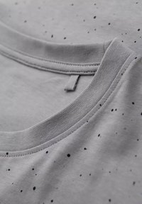 Camiseta de tela gris con un escote redondo, que presenta patrones moteados en negro y azul y una textura suave. Costura visible alrededor del cuello.