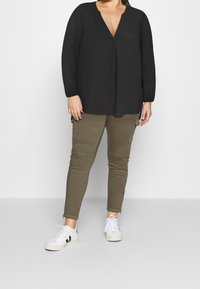 Blusa nera a maniche lunghe con scollatura a V profonda, abbinata a pantaloni cargo verde oliva con tasche laterali e sneakers bianche con dettagli neri.