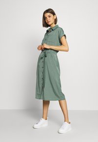 Abito verde a camicia con vita regolabile, maniche corte arrotolate, tasca frontale e orlo fino al ginocchio, abbinato a sneakers bianche.