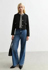 Veste croppée noire texturée avec deux poches avant et boutons argentés, portée sur un haut à rayures noir et blanc et un jean large bleu.
