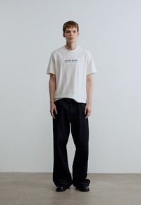 Λευκό βαμβακερό t-shirt με το κείμενο "Calvin Klein New York", συνδυασμένο με φαρδιά μαύρα τζιν παντελόνια και μαύρα επίπεδα παπούτσια. Απλός σχεδιασμός.